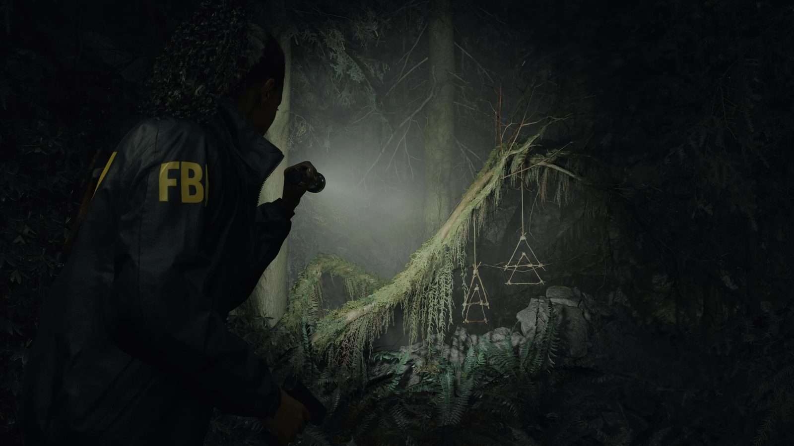 Saga Anderson, con chaqueta del FBI, ilumina con su linterna unos símbolos de ramitas que cuelgan de un árbol caído en un bosque de noche