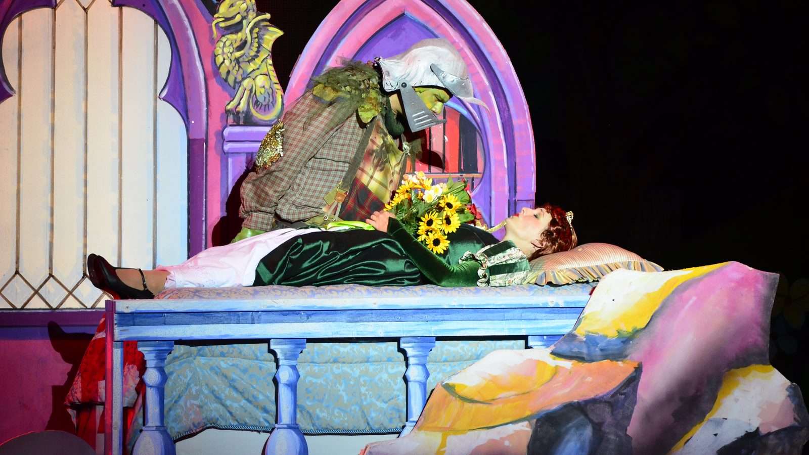 Escena de Shrek the Musical en un teatro: un actor con maquillaje de ogro verde y casco se inclina sobre una actriz con corona, recostada en una cama de cuento con un ramo de girasoles