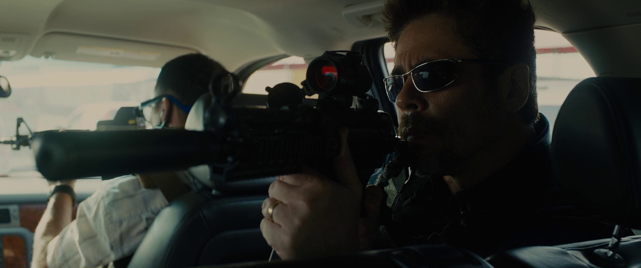 Benicio del Toro con gafas de sol sosteniendo un rifle con silenciador dentro de un automóvil en Sicario.