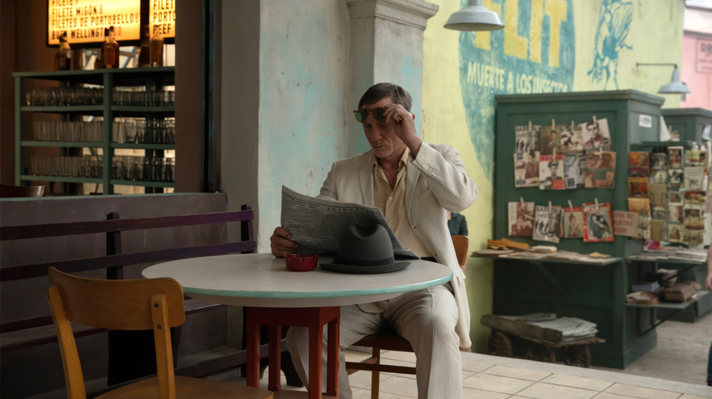 En Queer, Daniel Craig, vestido con un bonito traje de verano, se sienta en una mesa de café afuera de un restaurante y lee el periódico.