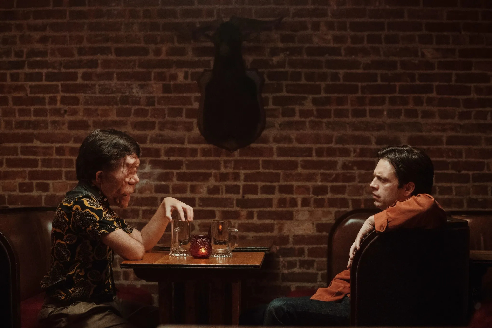 Adam Pearson y Sebastian Stan sentados uno frente al otro en un reservado de un restaurante en A Different Man.