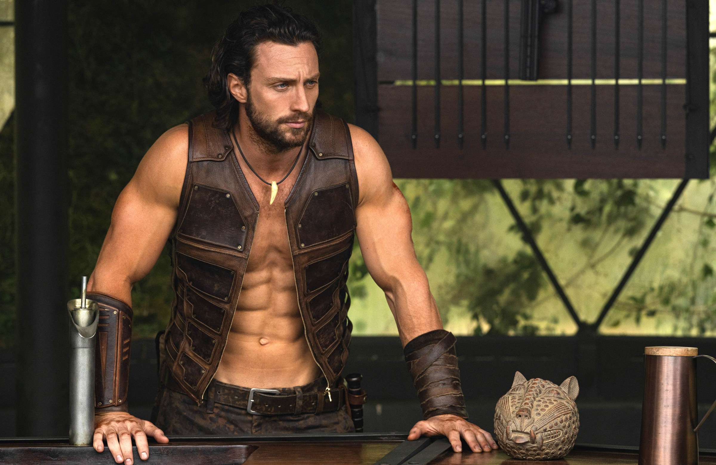 Kraven (Aaron Taylor-Johnson) está parado con las manos en una barandilla, vistiendo un chaleco de cuero abierto sin mangas y mostrando más abdominales de lo que parece humanamente posible en Kraven el Cazador.